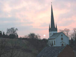 Das Foto zeigt die evangelische Pfarrkirche kurz vor Sonnenuntergang: eine helle Kirche im neuromanischem Stil mit spitzem Turm.