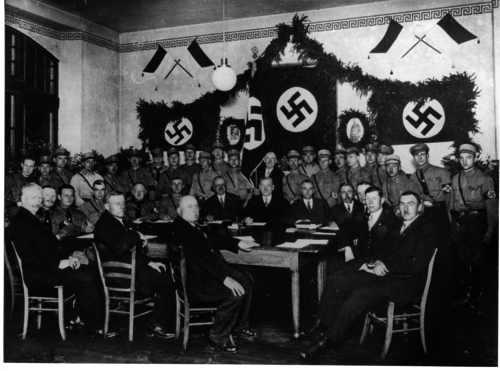 Abb. 8: Sitzung des Gemeinderats am 2. Mai 1933 im Turnsaal des Gemeindeschulhauses (heutiges Wilhelm-Spies-Haus). Lange Tischseite vorne mit Anzug von rechts nach links: SPD (Franz Rocker, Ludwig Hartmann, Lorenz Gabel) Kurze Tischseite links mit Uniform von links nach rechts: NSDAP (Anton Fleck II, Peter Mattes, Georg Boller, Hermann Koch, Philipp Hecker) Lange Tischseite hinten mit Anzug von links nach rechts: Beigeordneter Johann Schweibächer, Bürgermeister Jakob Spies V, Gemeindesekretär Adam Oehrlein Kurze Tischseite rechts mit Anzug von links nach rechts: Zentrum (Andreas Heftrich, Heinrich Schreiner, Josef Schon II., Josef A. Görlich II) Hinter den Mitgliedern des Gemeinderats sind die im Durchschnitt jüngeren Mitglieder der SA versammelt.