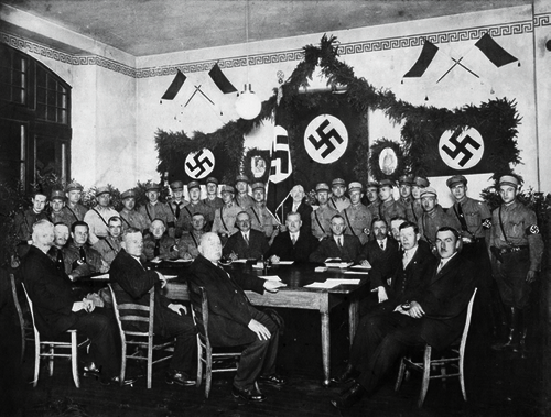 Abb. 8: Sitzung des Gemeinderats am 2. Mai 1933 im Turnsaal des Gemeindeschulhauses (heutiges Wilhelm-Spies-Haus). Lange Tischseite vorne mit Anzug von rechts nach links: SPD (Franz Rocker, Ludwig Hartmann, Lorenz Gabel) Kurze Tischseite links mit Uniform von links nach rechts: NSDAP (Anton Fleck II, Peter Mattes, Georg Boller, Hermann Koch, Philipp Hecker) Lange Tischseite hinten mit Anzug von links nach rechts: Beigeordneter Johann Schweibächer, Bürgermeister Jakob Spies V, Gemeindesekretär Adam Oehrlein Kurze Tischseite rechts mit Anzug von links nach rechts: Zentrum (Andreas Heftrich, Heinrich Schreiner, Josef Schon II., Josef A. Görlich II) Hinter den Mitgliedern des Gemeinderats sind die im Durchschnitt jüngeren Mitglieder der SA versammelt.