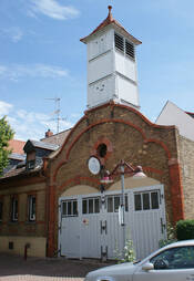 Das Bild zeigt das Alte Spritzenhaus in Bretzenheim: Ein Backsteinhaus mit zwei Toren für Fahrzeuge und Löschgeräte. Darüber sieht man den hölzernen Turm fürs Aufhängen der Schläuche.