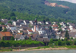 Auf dem Foto sieht man den Ortskern von Trechtingshausen von der anderen Rheinseite aus aufgenommen. Zu erkennen sind die Pfarrkirche und viele Häuser. Im Vordergrund ist noch knapp der Rhein zu sehen.